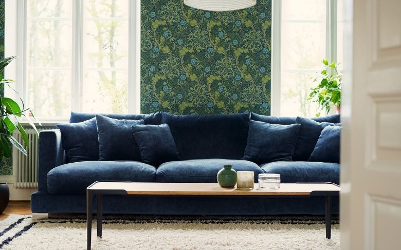Canapé déhoussable,Calvi, 4 places en velours bleu marine. Disposé devant un mur au papier peint vert fleuri et face à une table basse en bois aux pieds métalliques. 